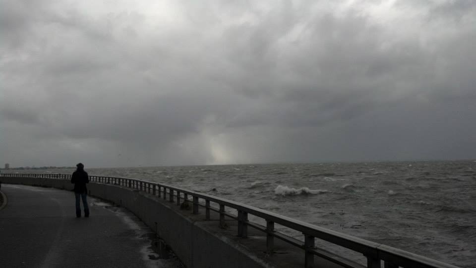This photo was taken by the Verrazzano bridge the day after Hurricane Sandy. A person stands on the promenade looking out over the dark, rolling ocean. The sky is grey and stormy above.